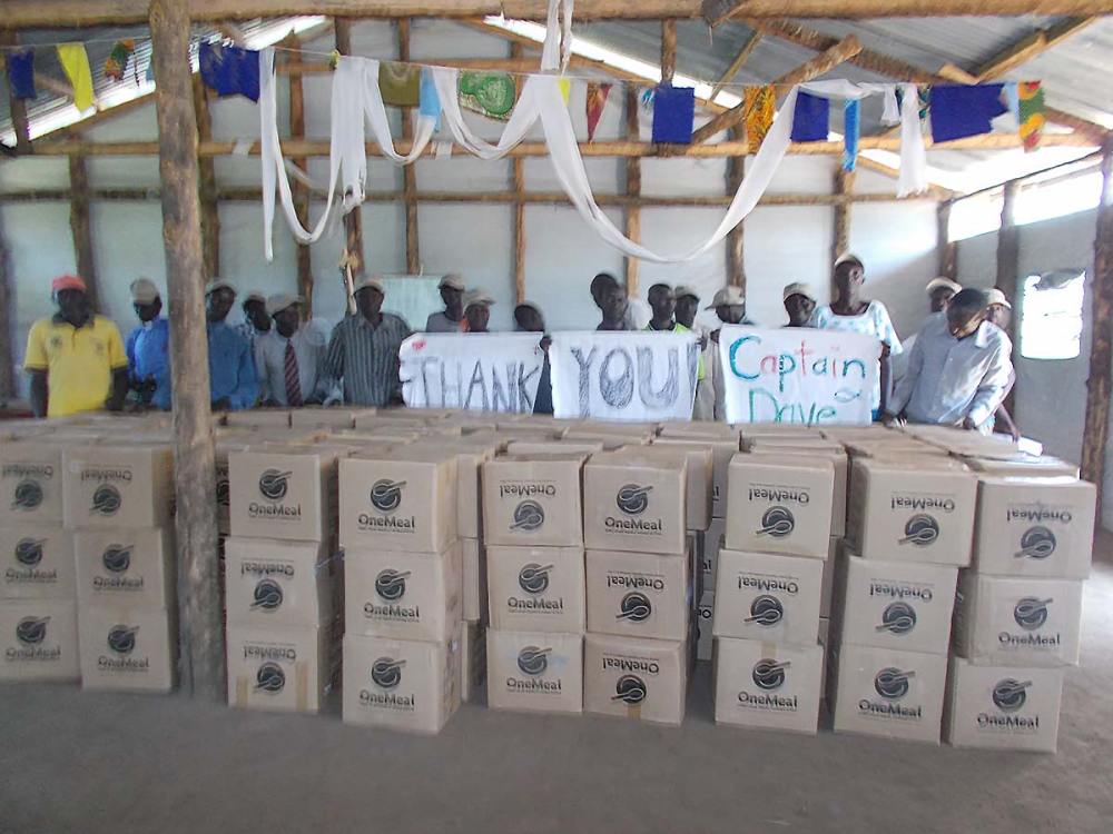 Ugandan refugees hold sign Thank You Capt Dave while standing behind OneMeal boxes of packed food.